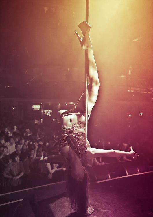 woman performing on stage in front of audience in heels doing leg hang, pole fitness, pole dancing